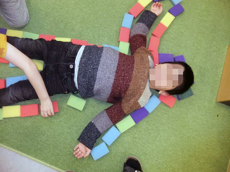 Kind liegt mit gespreizten Armen am Boden, die Körperform ist mit kleinen Schwämmen umrandet.