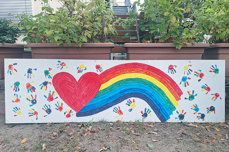 großes Plakat mit vielen bunten Abdrücken von Kinderhänden, in der Mitte ein Regenbogen und ein Herz