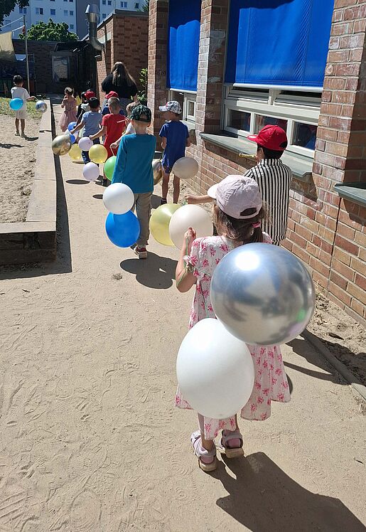 Kinder mit Ballons und Tröten ziehen über den Kita-Platz.