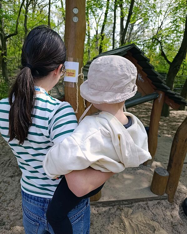 Frau mit Kind auf dem Arm vor einem "Schattenplatz"-Schild