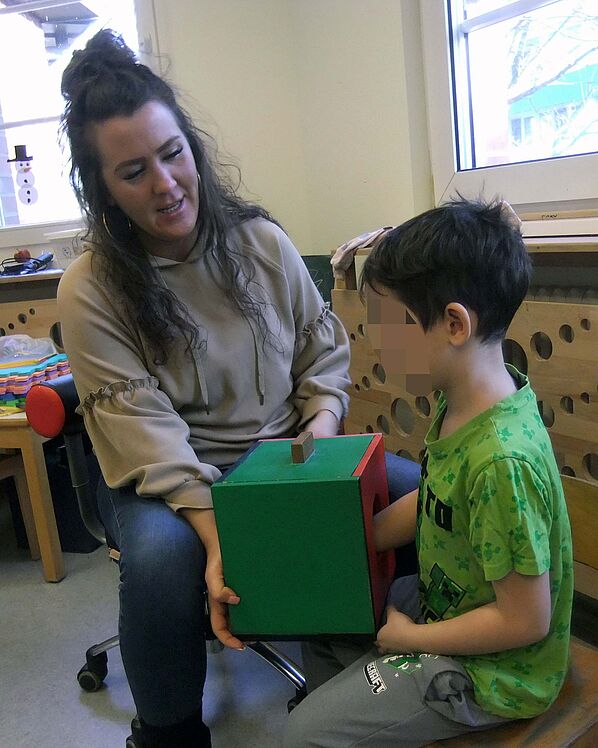 Kind mit kurzen dunklen Haaren steckt Hand in Kasten, um Formen zu ertasten, der Kasten wird von der Erzieherin gehalten.