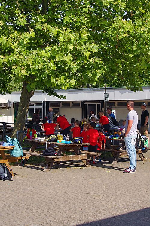 Kinder und Erwachsene bei Snacks und Getränken im Schatten unter einem Baum vor dem Vereinshaus