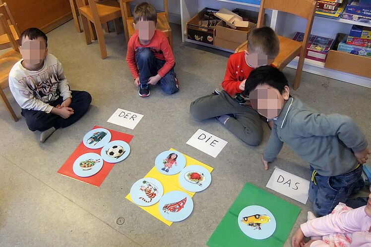 Auf dem Boden sitzende Kinder betrachtet Kärtchen mit Figuren wie Autos etc. vor sich, denen die jeweils dem passenden Artikel (der, die, das) zugeordnet sind.