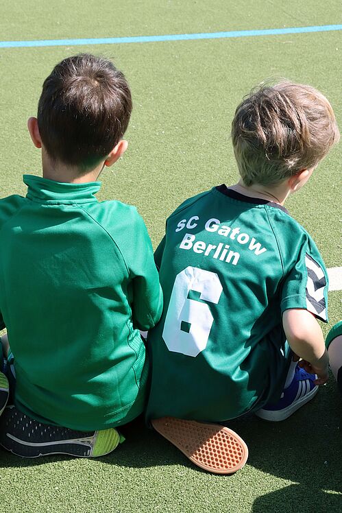 Zwei Kinder in grünen Trikots (von hinten) sitzen auf dem Platz