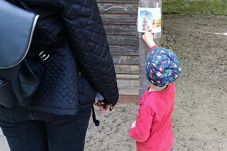 Kind zeigt erwachsener Person das Schild "Hier ist ein Schattenplatz"