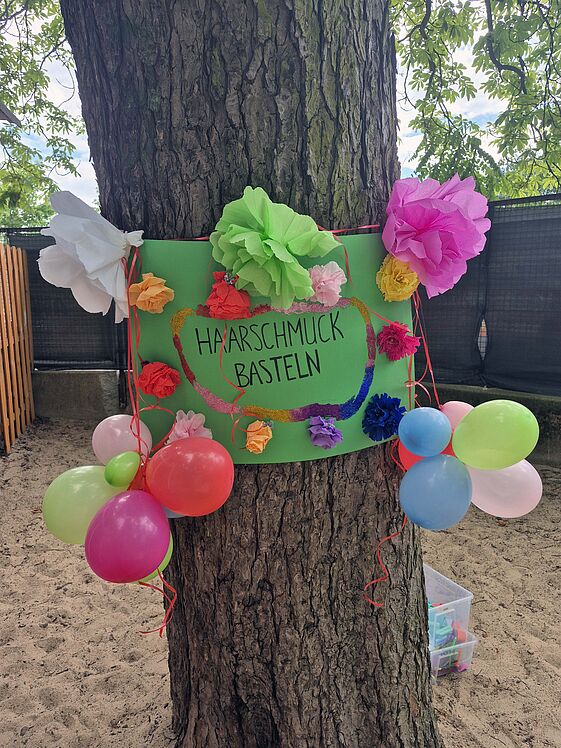 An einem Baumstamm befestigtes Plakat zur Aktion "Haarschmuck basteln", verziehrt mit Papierblumen und Luftballons
