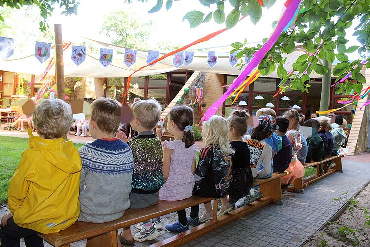 Rückansicht von auf einer Bank sitzenden Kindern im geschmückten Außenbereich der Kita.
