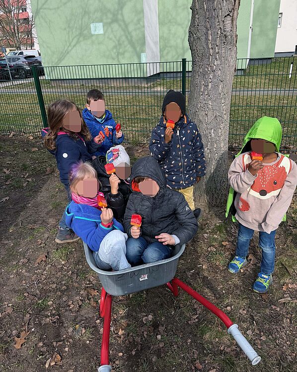 Ein paar Kinder sitzen in einer Schubkarre, andere stehen daneben, alle schlecken ein Eis.