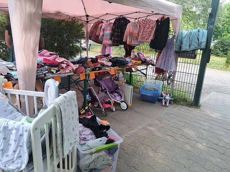 Flohmarktstand mit Kleidung, im Vordergrund ist der Teil eines Gitterbetts mit Ausstattung zu sehen.